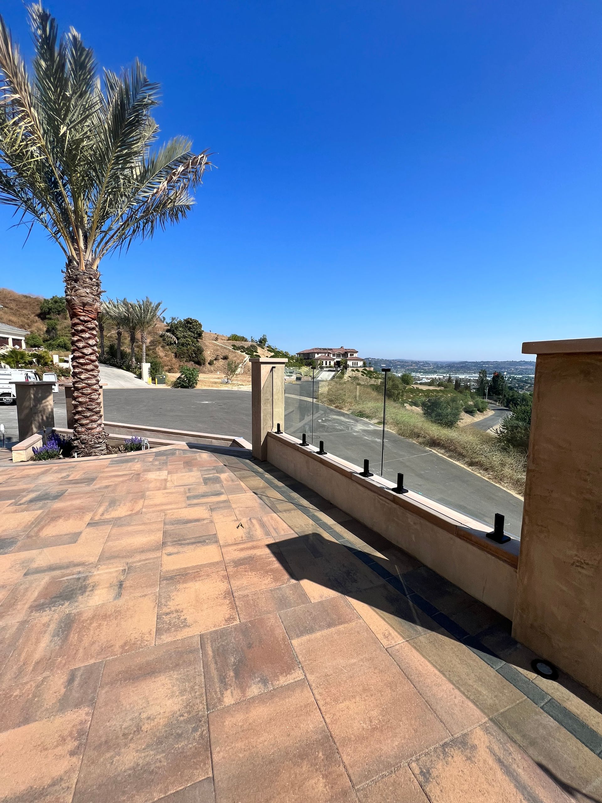 Patio with glass railing overlooking a street and distant landscape, under a clear blue sky.