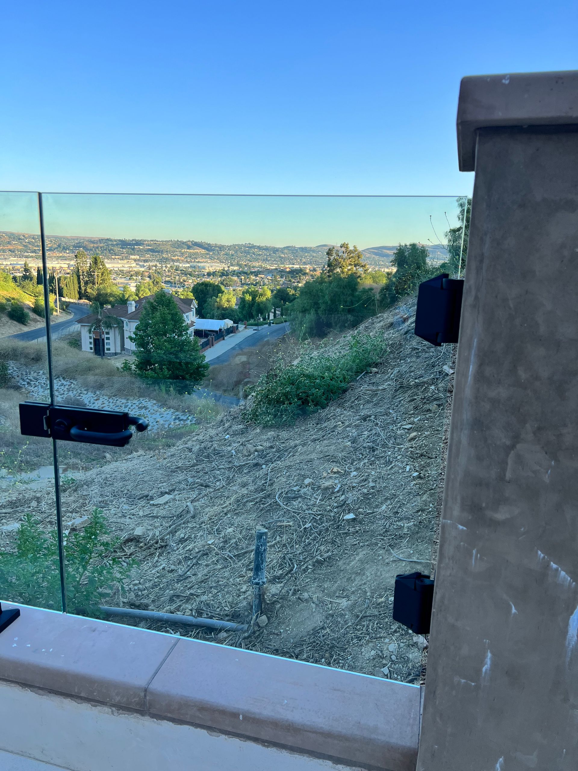 Glass railing on a patio overlooking a hillside with a distant city under a blue sky.