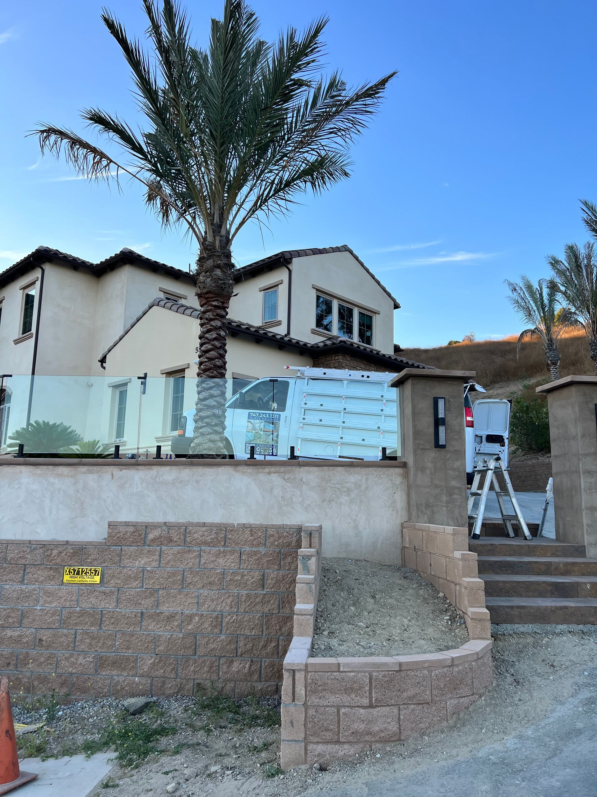 Beige house behind a glass railing with a palm tree. Brick wall and stairs in foreground.
