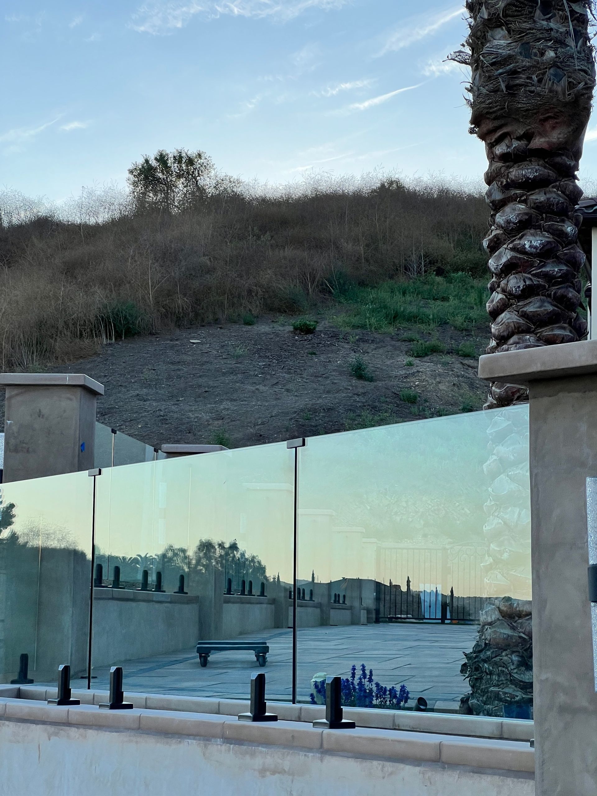 Glass fence with black posts, concrete base, with a hilly landscape in the background, under a blue sky.