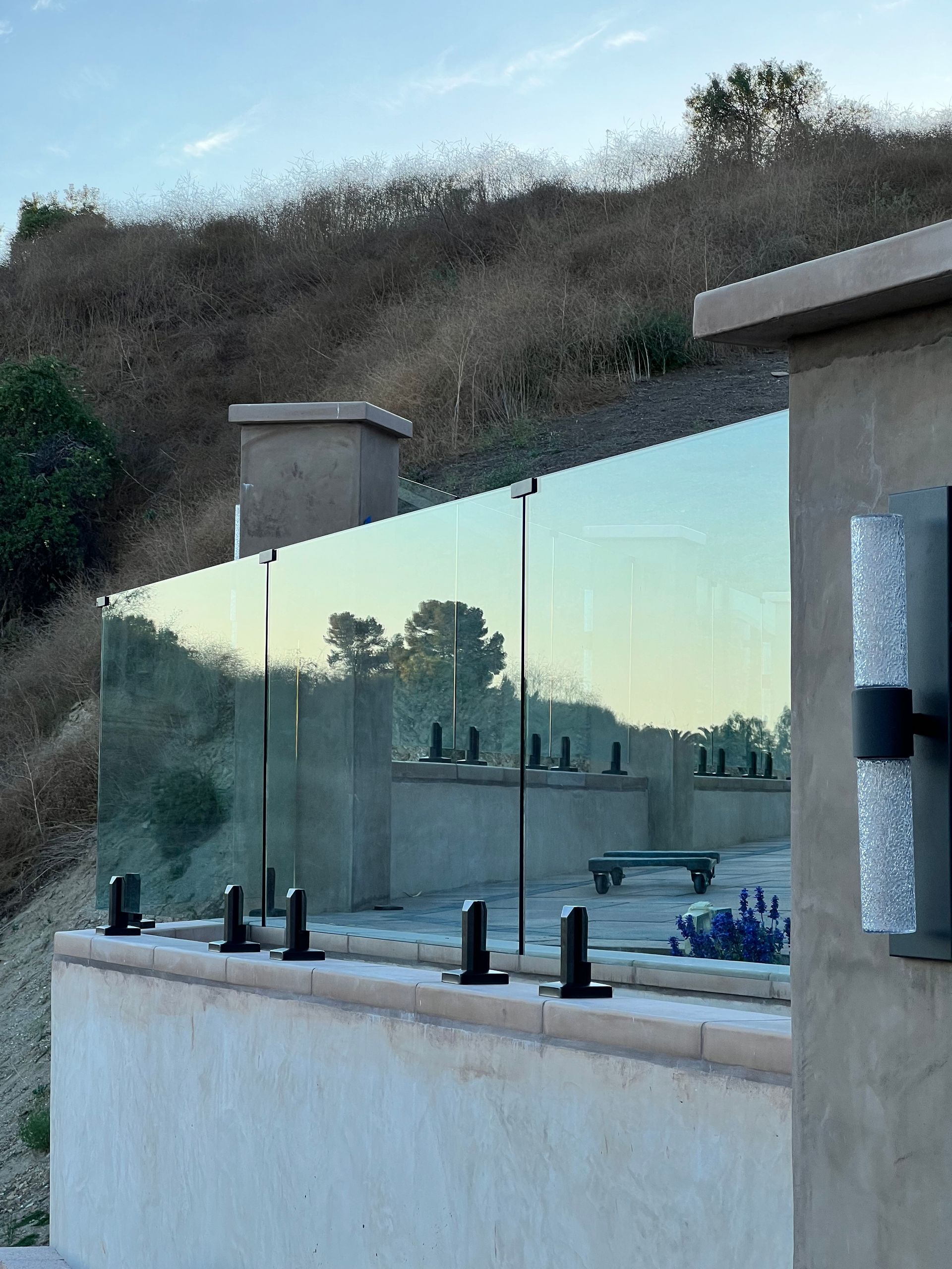 Glass railing on a patio overlooking a hillside; black hardware and a decorative wall sconce are visible.