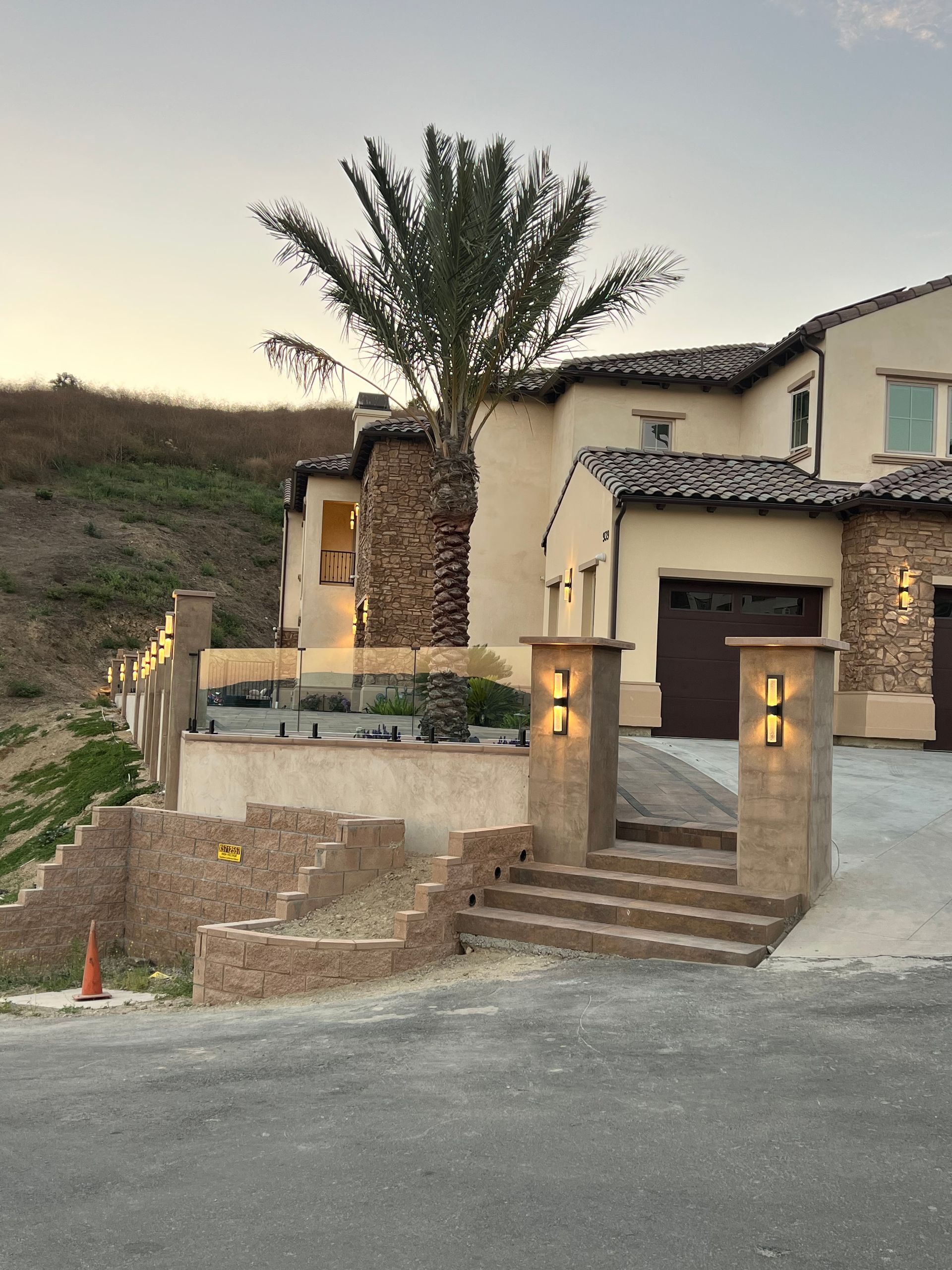 Beige stucco home with stone accents, palm tree, and steps leading to the entrance. Dusk lighting.