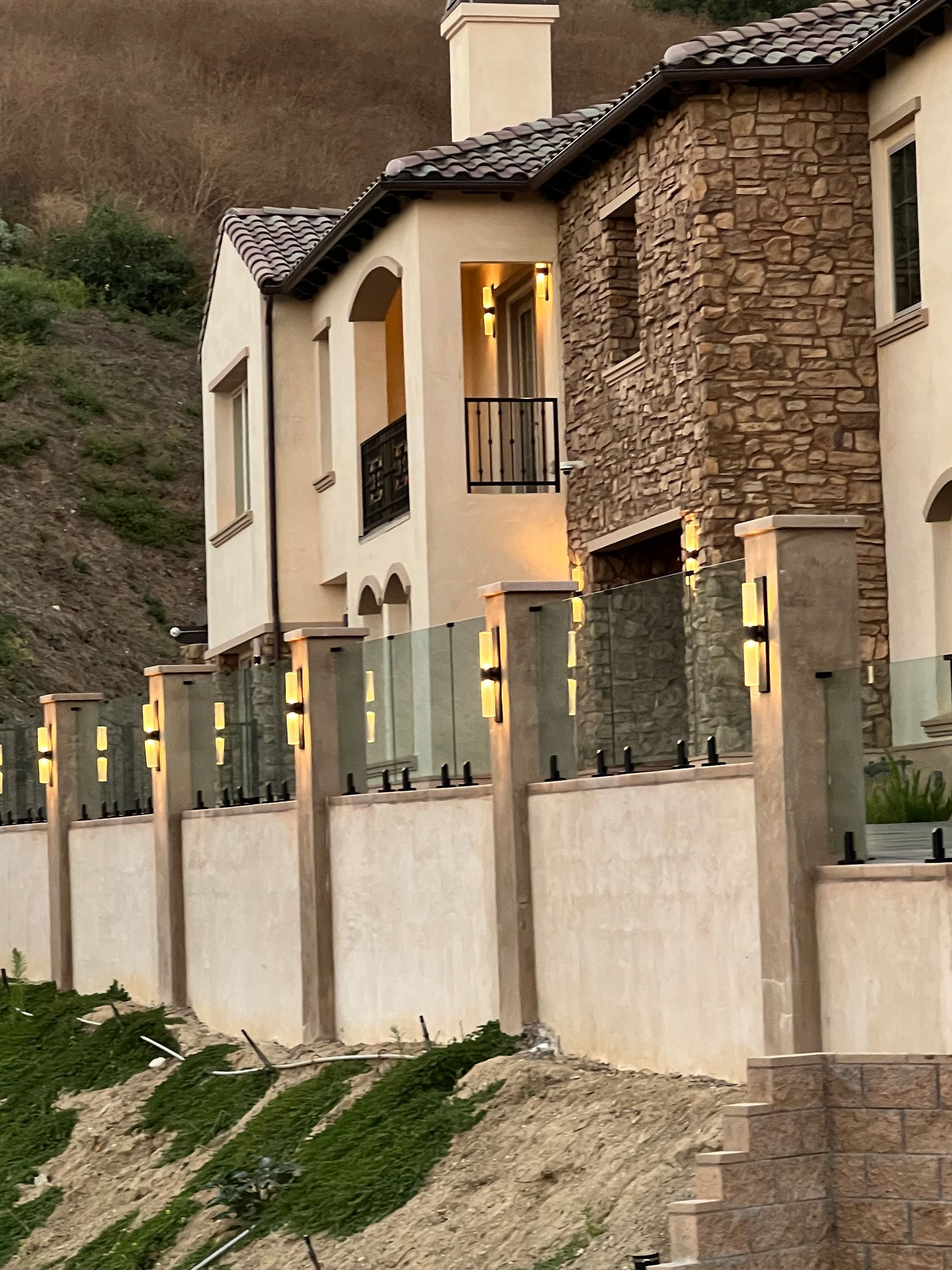 Beige and stone home with a glass and concrete fence, against a hillside with green plants.