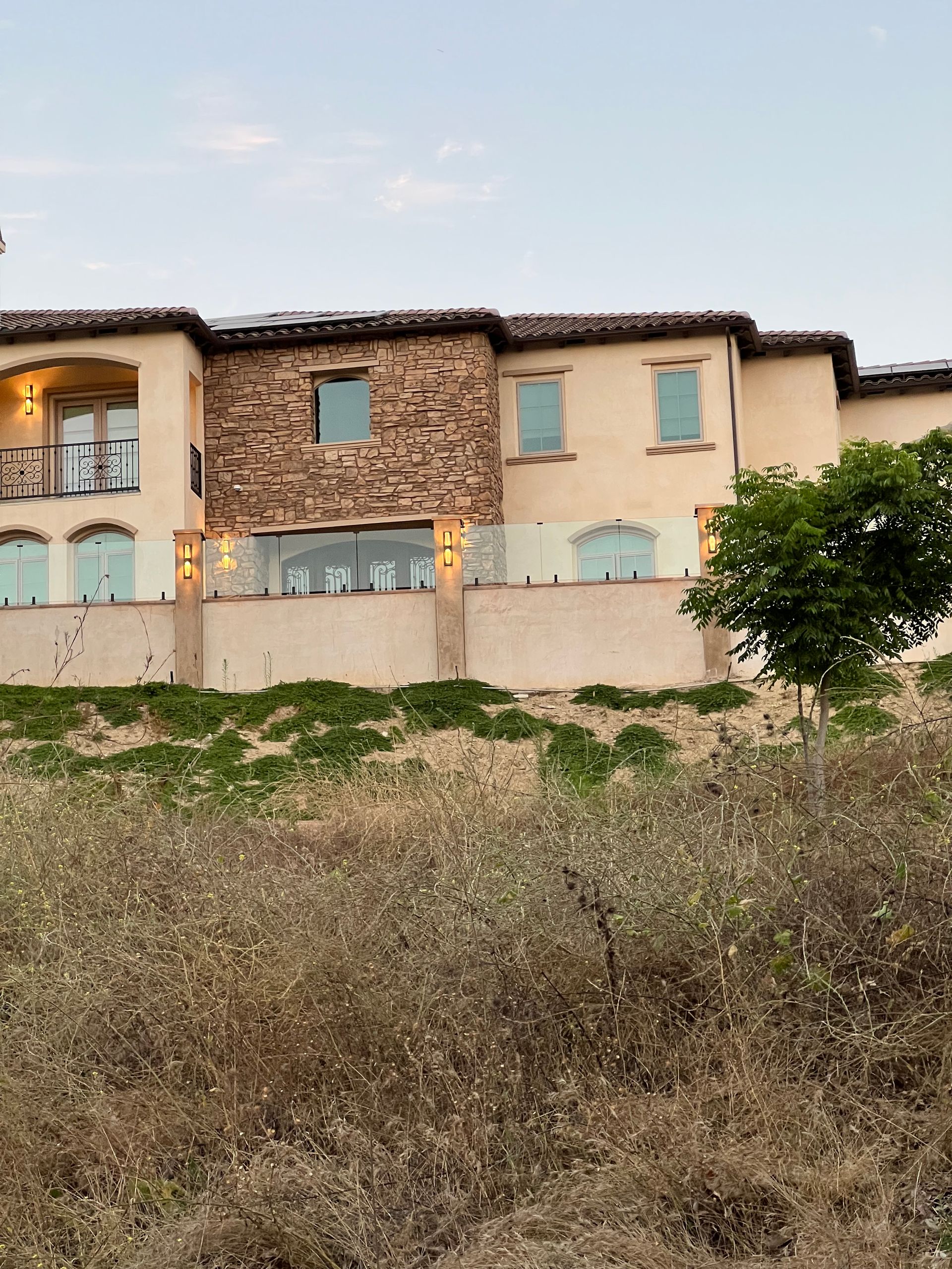 Two-story building with tan stucco and stone facade, overlooking overgrown vegetation, under a pale sky.