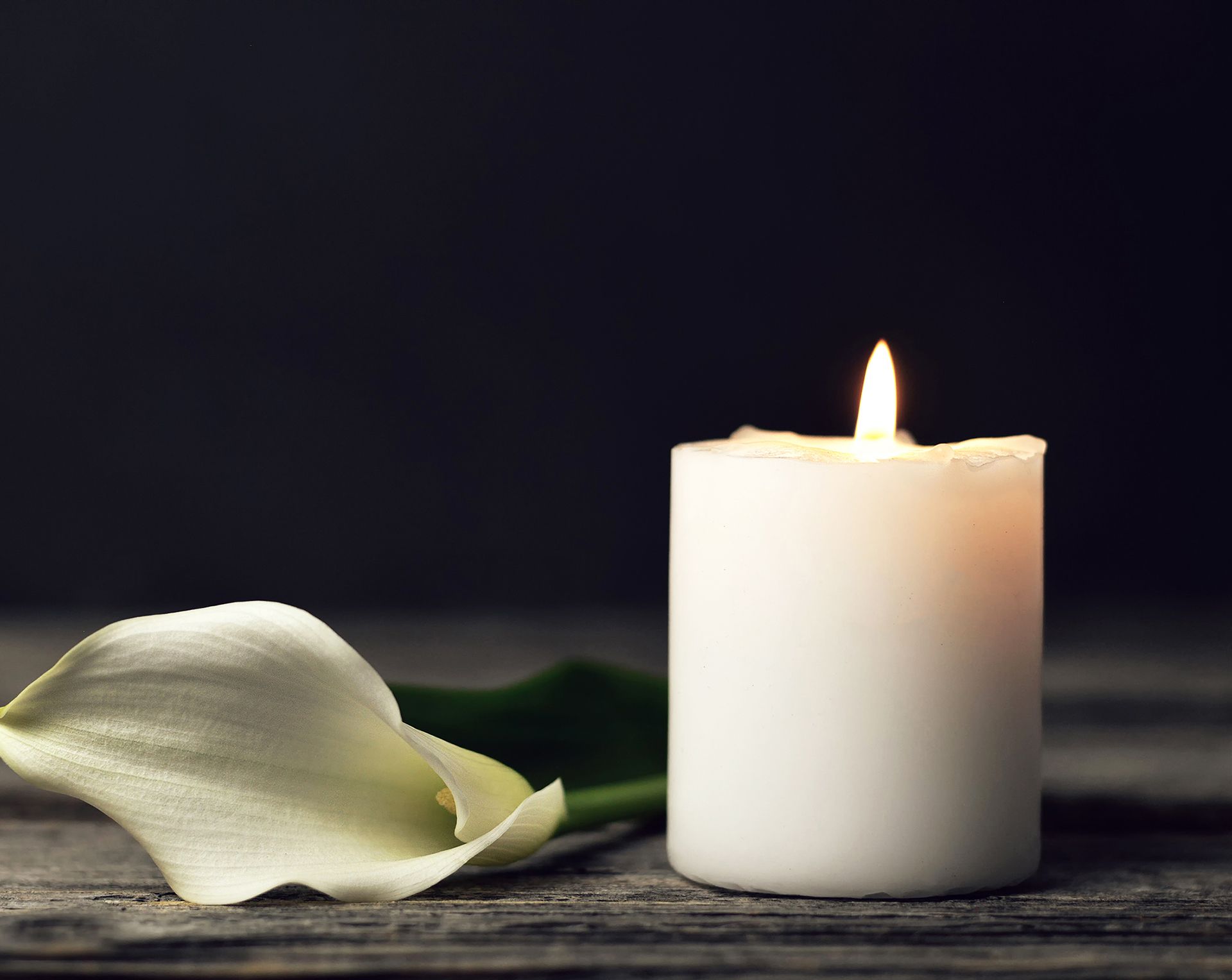 A white candle is lit next to a white flower on a wooden table.
