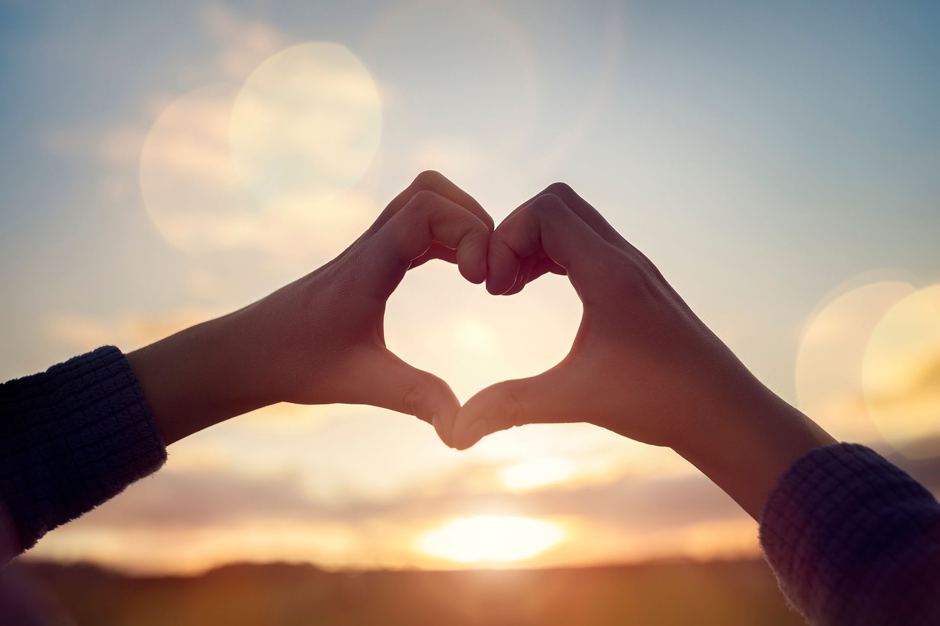 A person is making a heart shape with their hands at sunset.