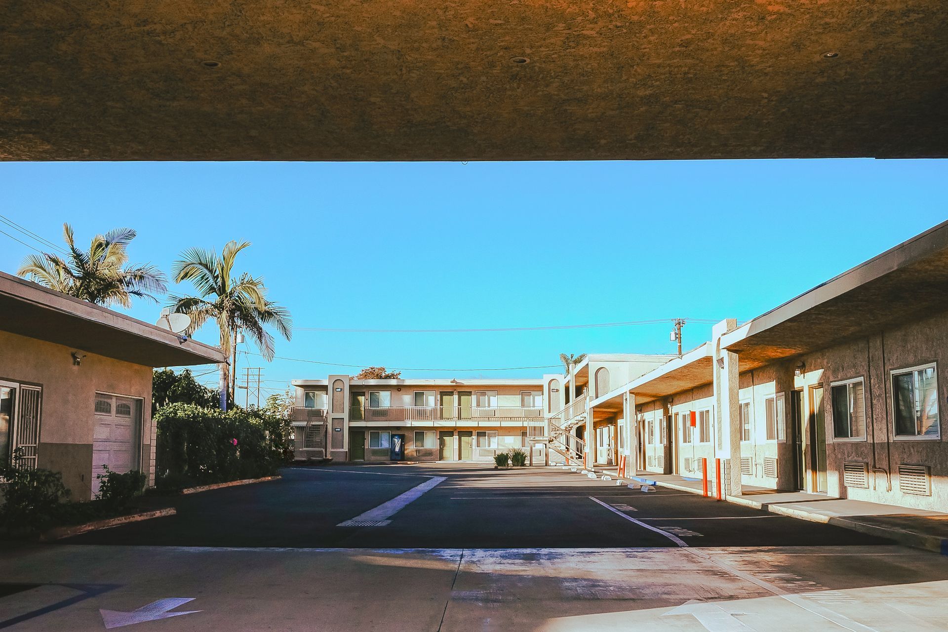 Motel with palm trees, blue sky. Concrete structures, asphalt parking lot.