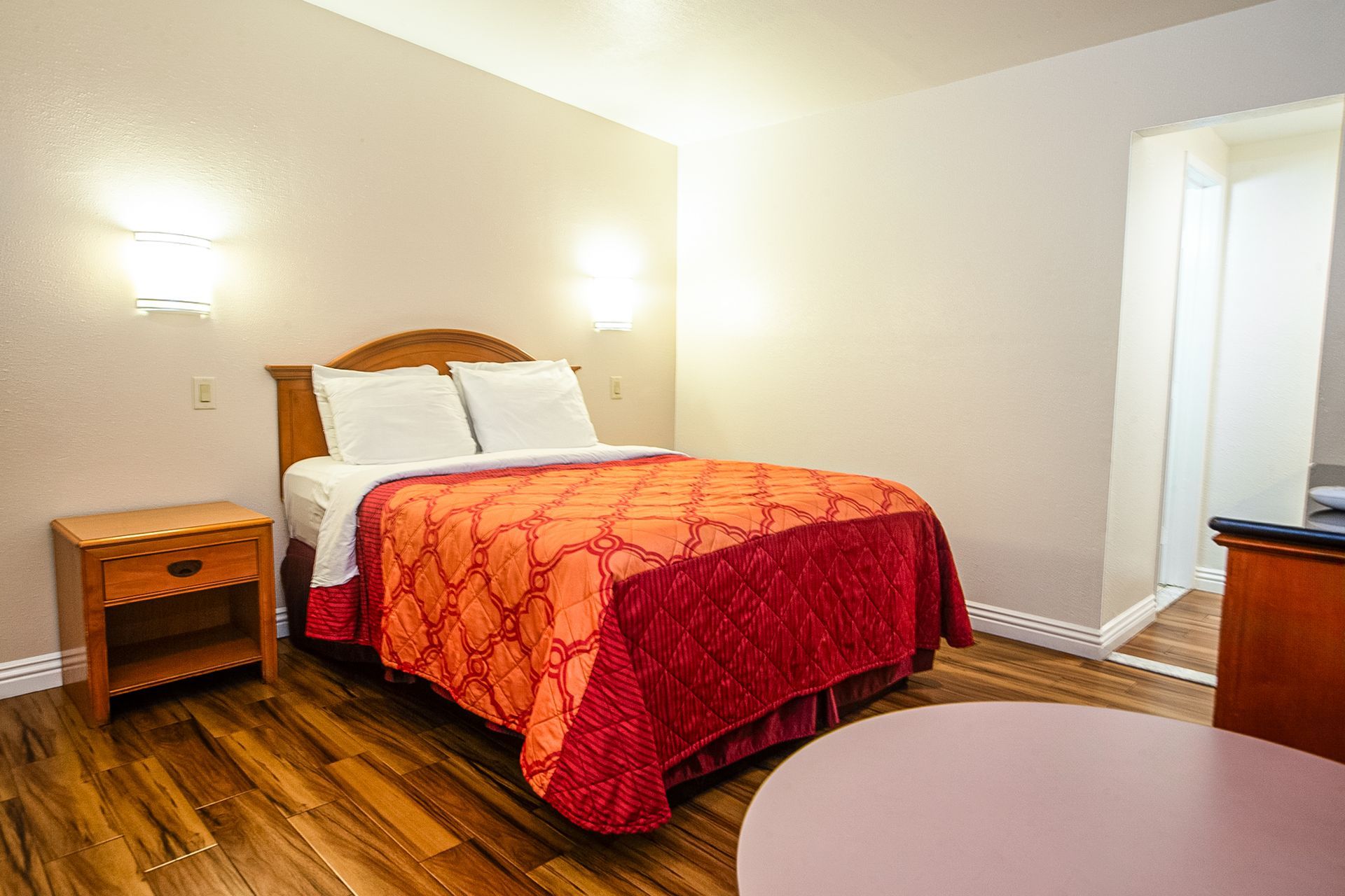 Hotel room with a bed, nightstand, and small table. Wooden floor and orange/red bedspread.