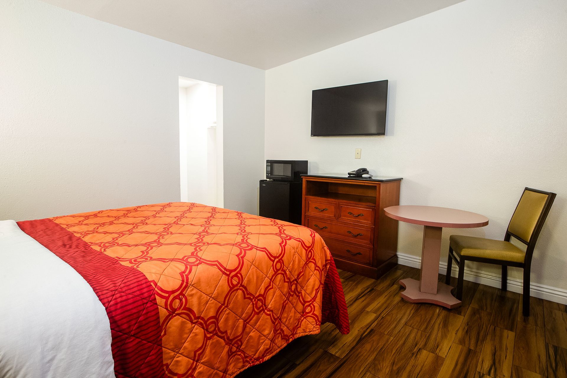 Hotel room with bed, TV, table, and microwave. Orange bedspread, wooden floor, and a small refrigerator.