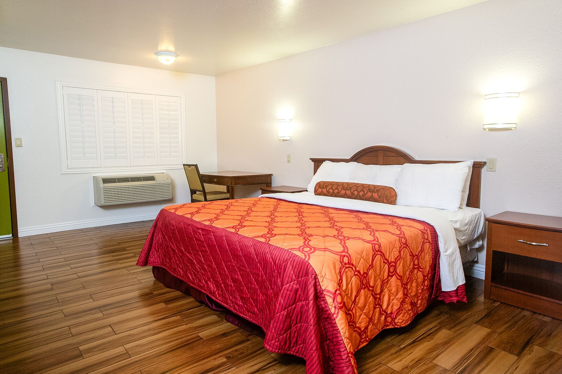 Bedroom with king bed, orange quilt, wooden floor, desk, air conditioner, and nightstand.