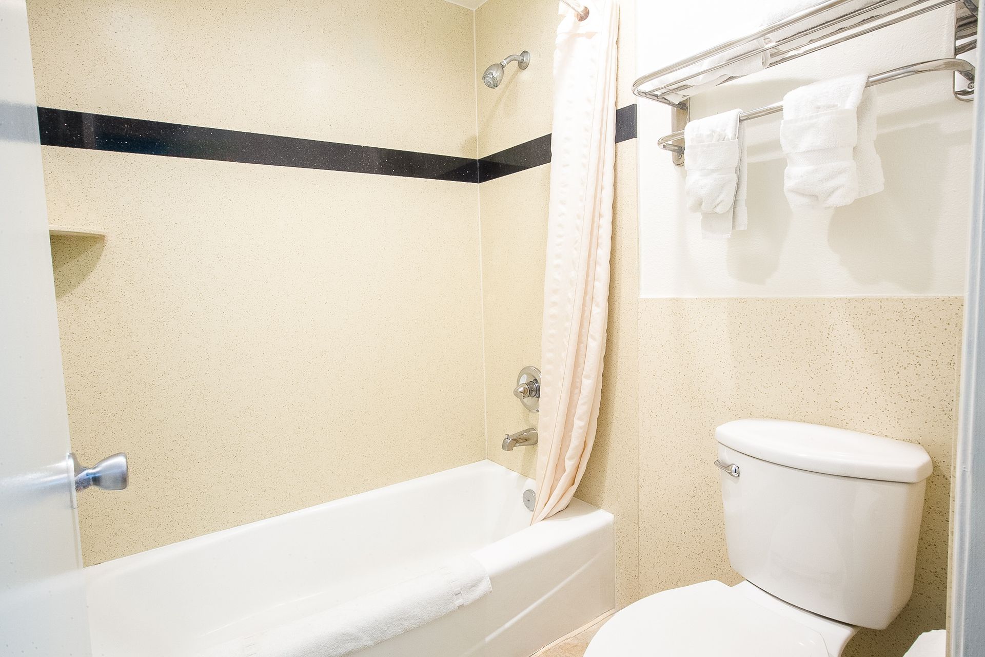 Bathroom with bathtub, toilet, and towel rack. Beige speckled walls, black stripe, white towels, and a shower curtain.