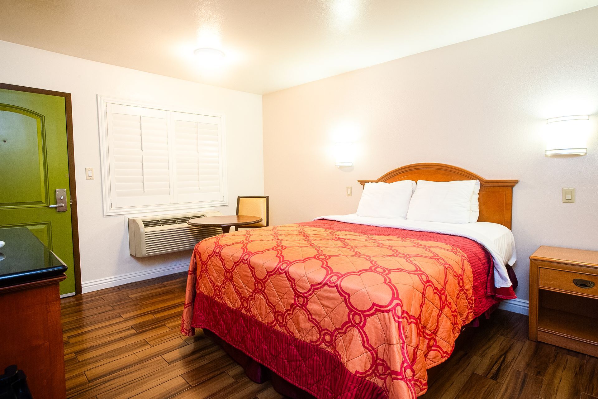 Hotel room with a bed, nightstand, window, and green door. Orange patterned bedspread.