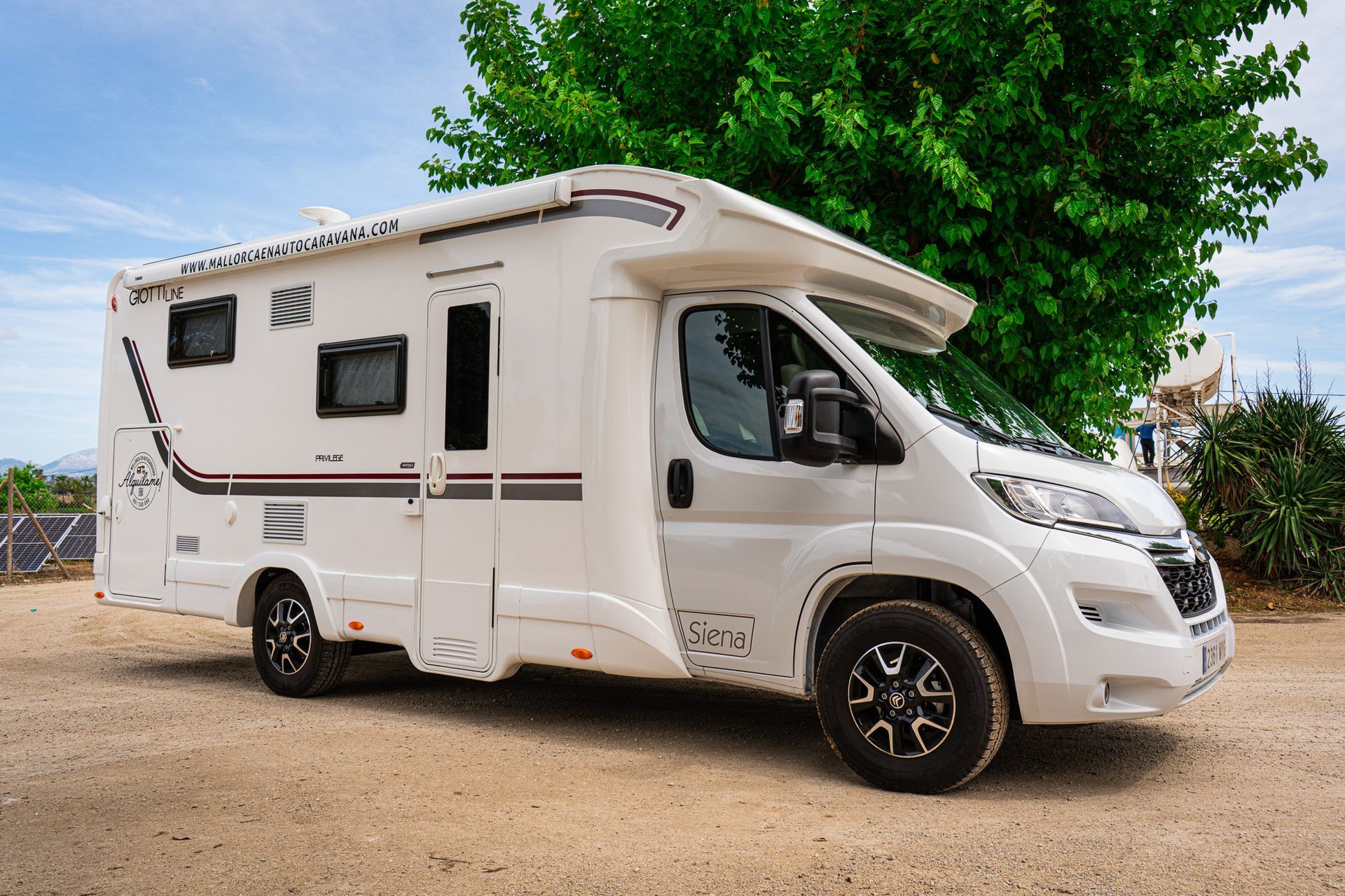 Una casa rodante blanca está estacionada en un lote de grava al lado de un árbol.