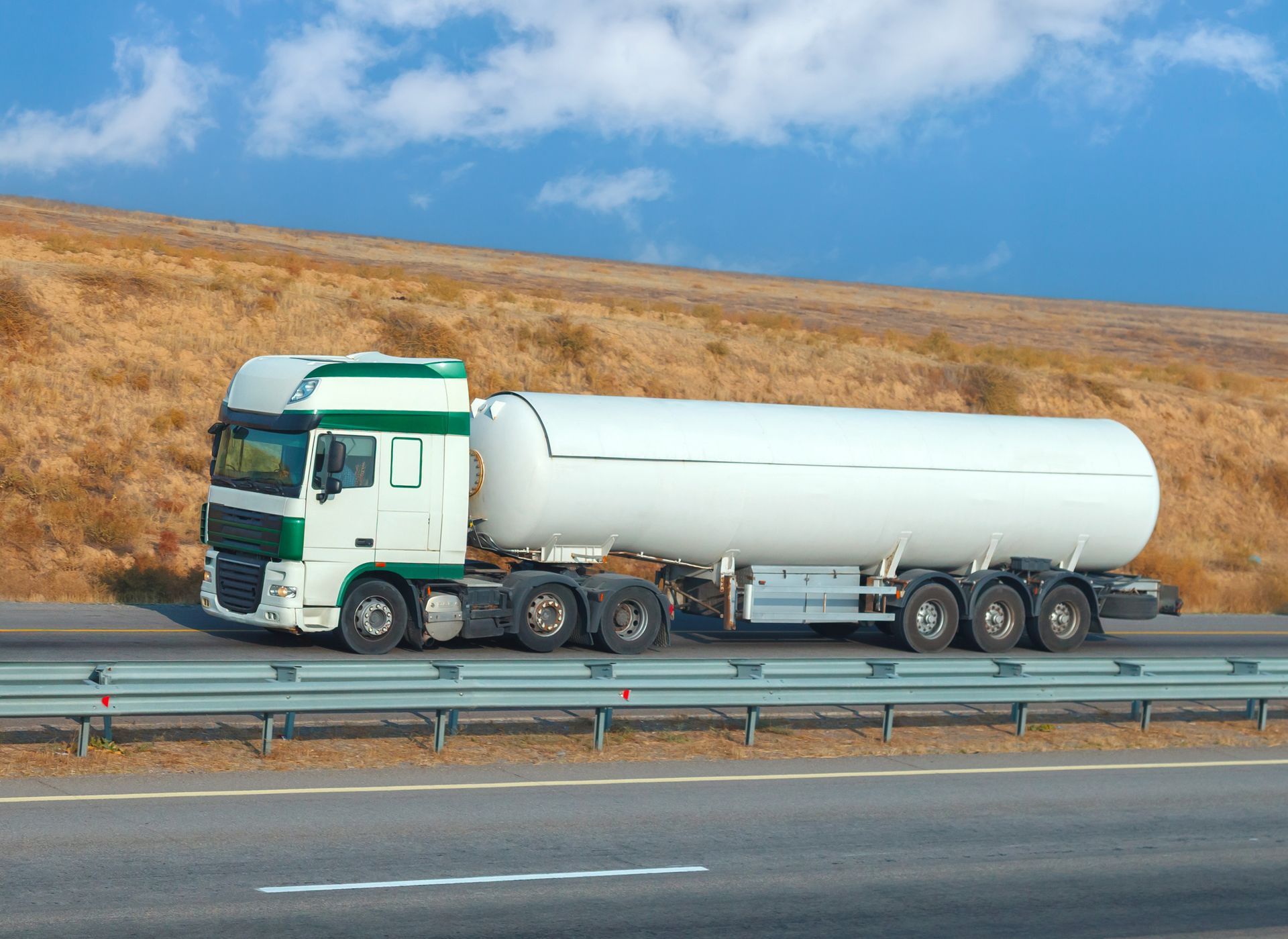 A large white gas tanker truck driving along the roadway beneath a bright blue sky.