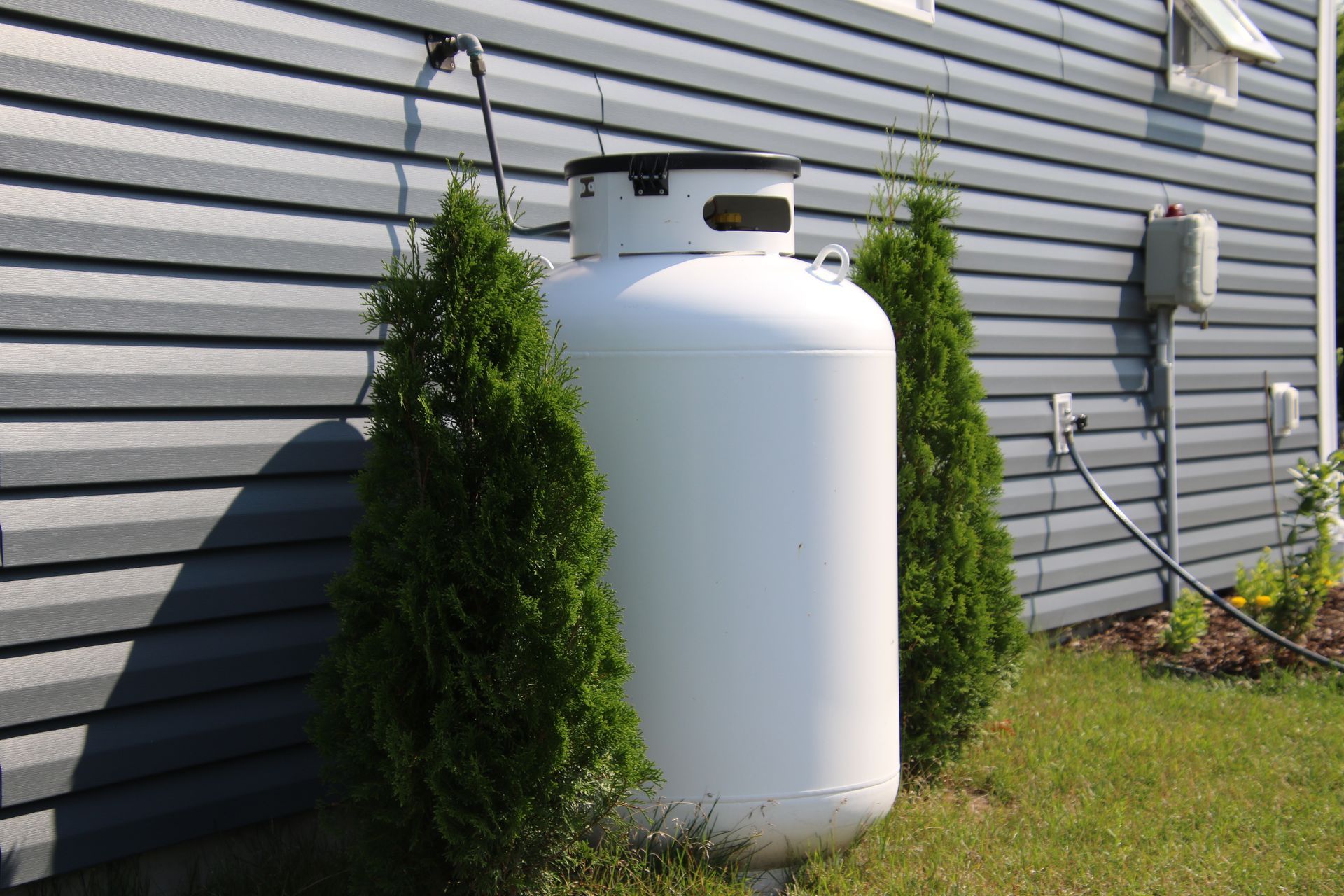 Outdoor propane tank beside house siding, flanked by small evergreen shrubs. Outdoor propane tank beside house siding, flanked by small evergreen shrubs.