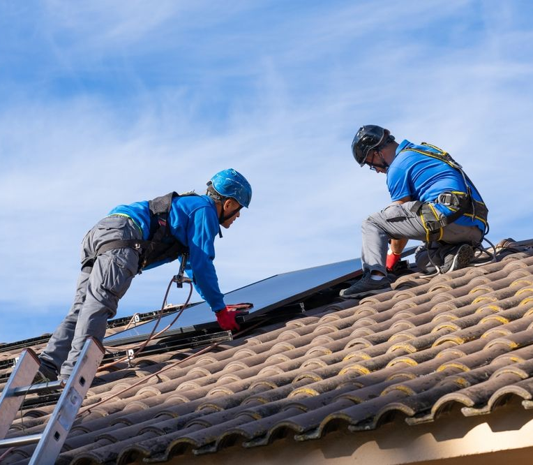 Due operai in tuta di sicurezza installano pannelli solari su un tetto piastrellato sotto un cielo azzurro.