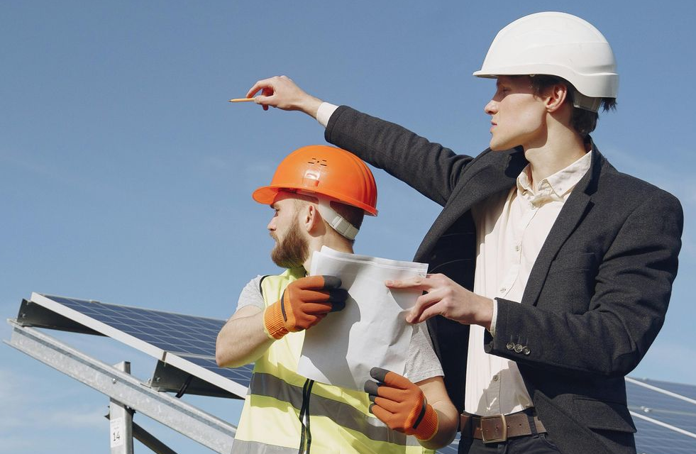 Due uomini con il casco vicino ai pannelli solari; uno indica, l'altro mostra i piani.