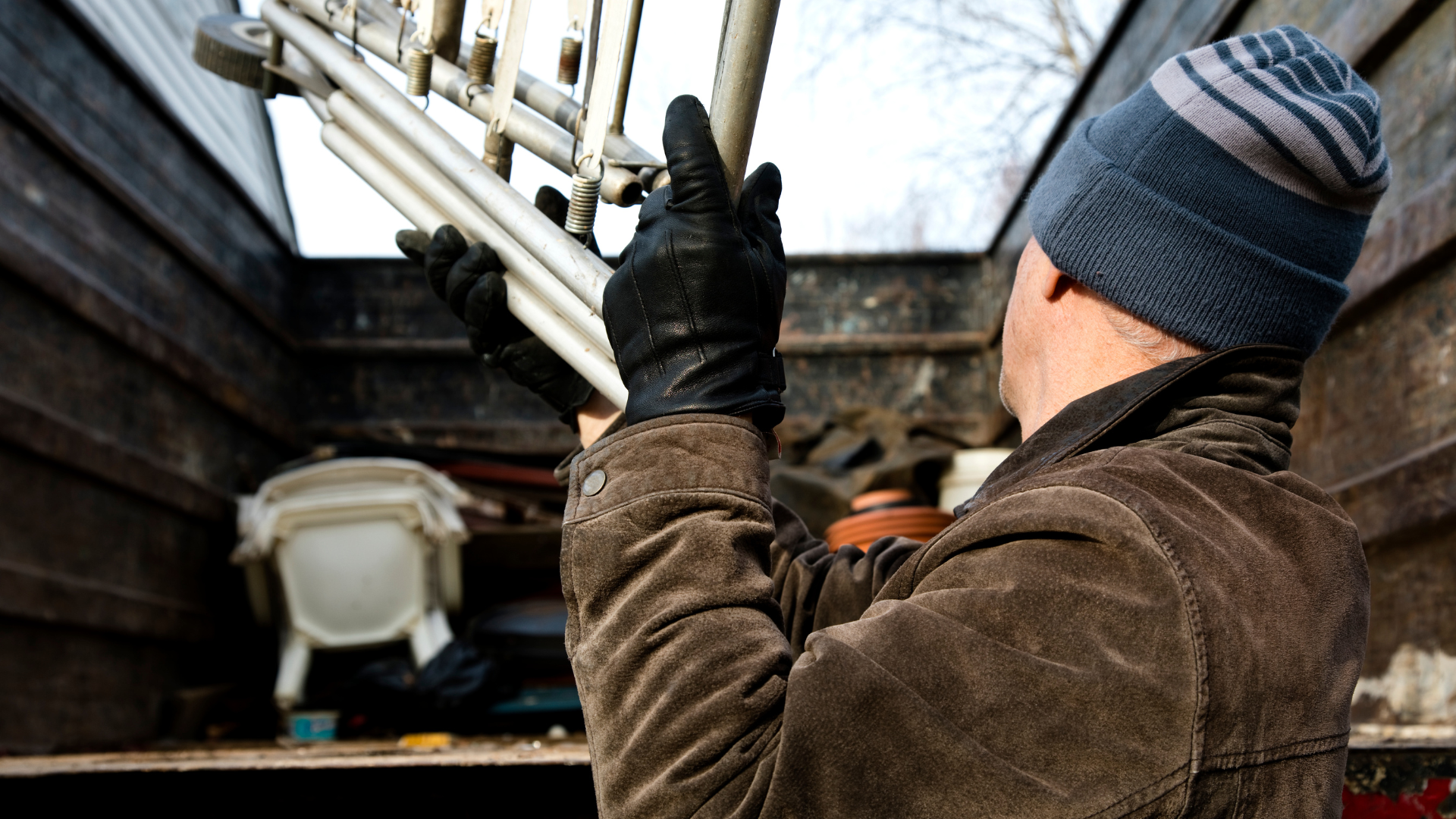 A man wearing gloves and a hat is working on a light fixture in the back of a truck.