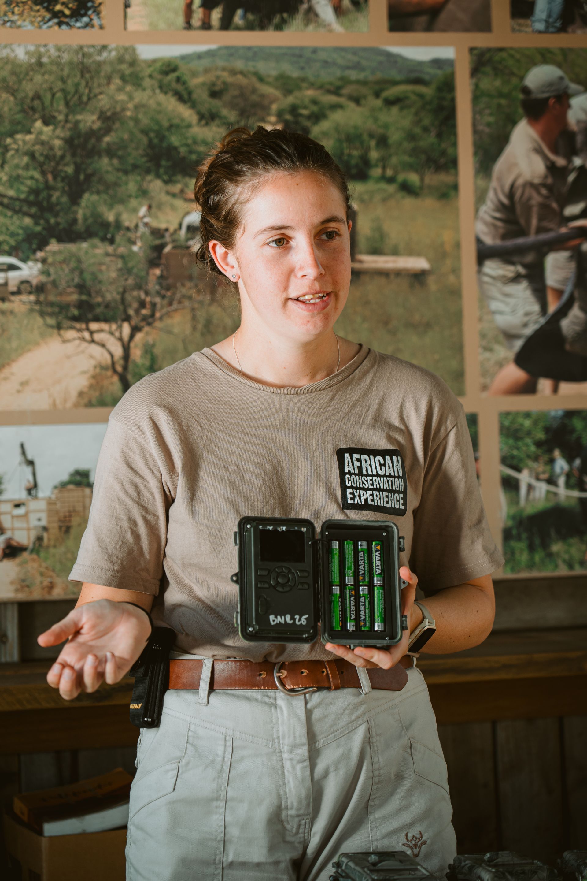 Woman in tan shirt holding camera trap, gesturing. Background includes nature photos and a person in khaki.