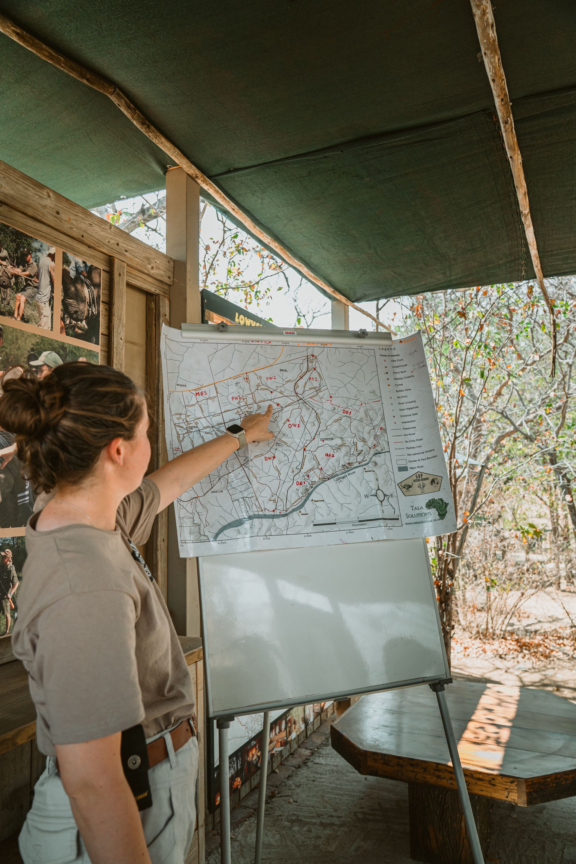 Woman points at a map on a whiteboard outdoors, under a shaded shelter.