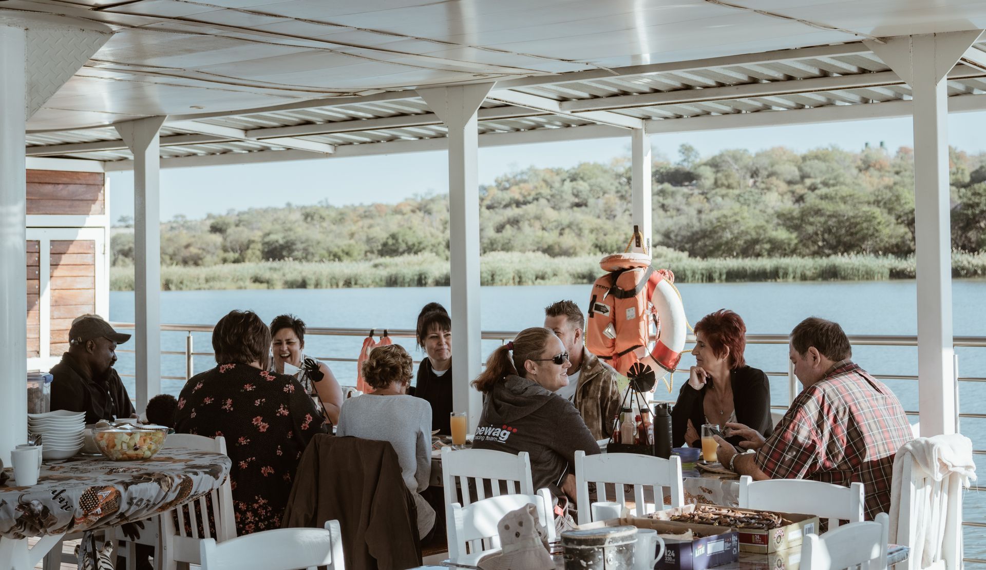 People seated at tables on a boat deck, enjoying a meal, with a waterway and trees in the background.
