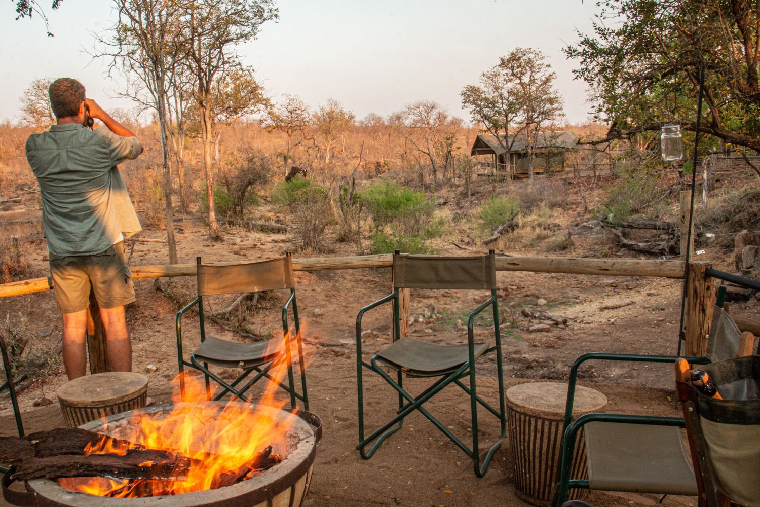 Man looking through binoculars the wildlife from a campsite with a fire and chairs.