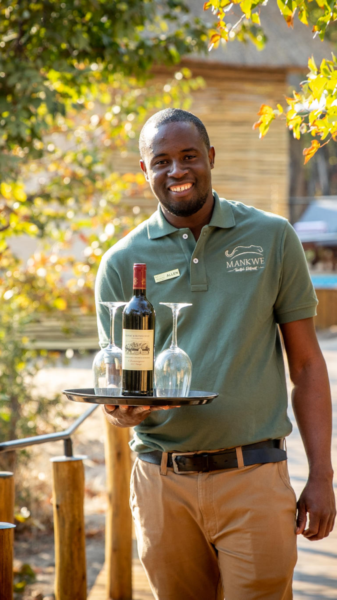 Waiter smiling, holding tray with wine bottle and glasses outdoors, wearing green shirt and khaki pants.