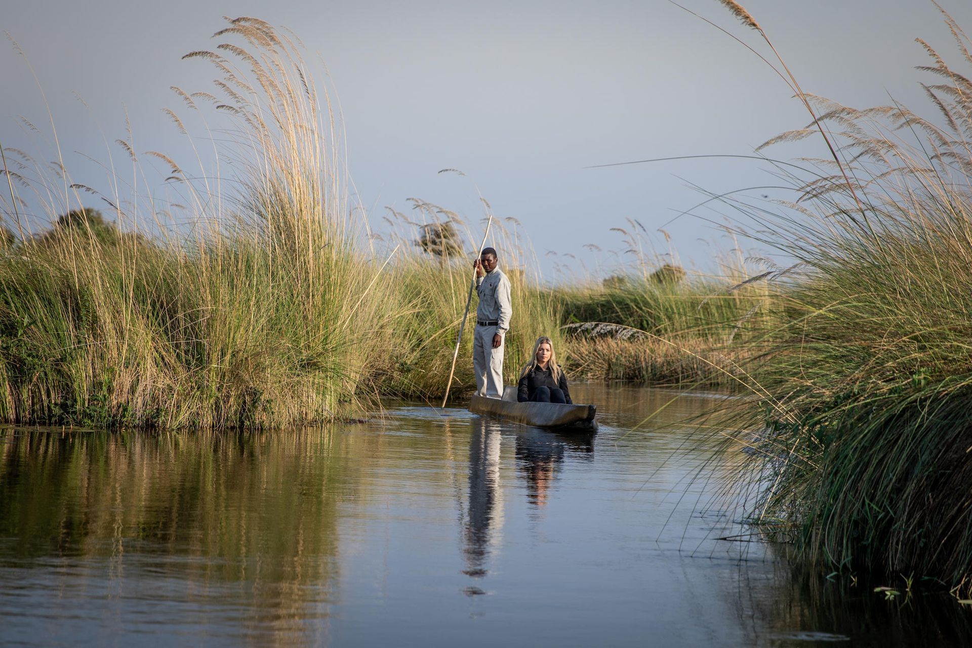 Two people in a wooden canoe navigate a calm river, surrounded by tall grasses, under a clear sky.
