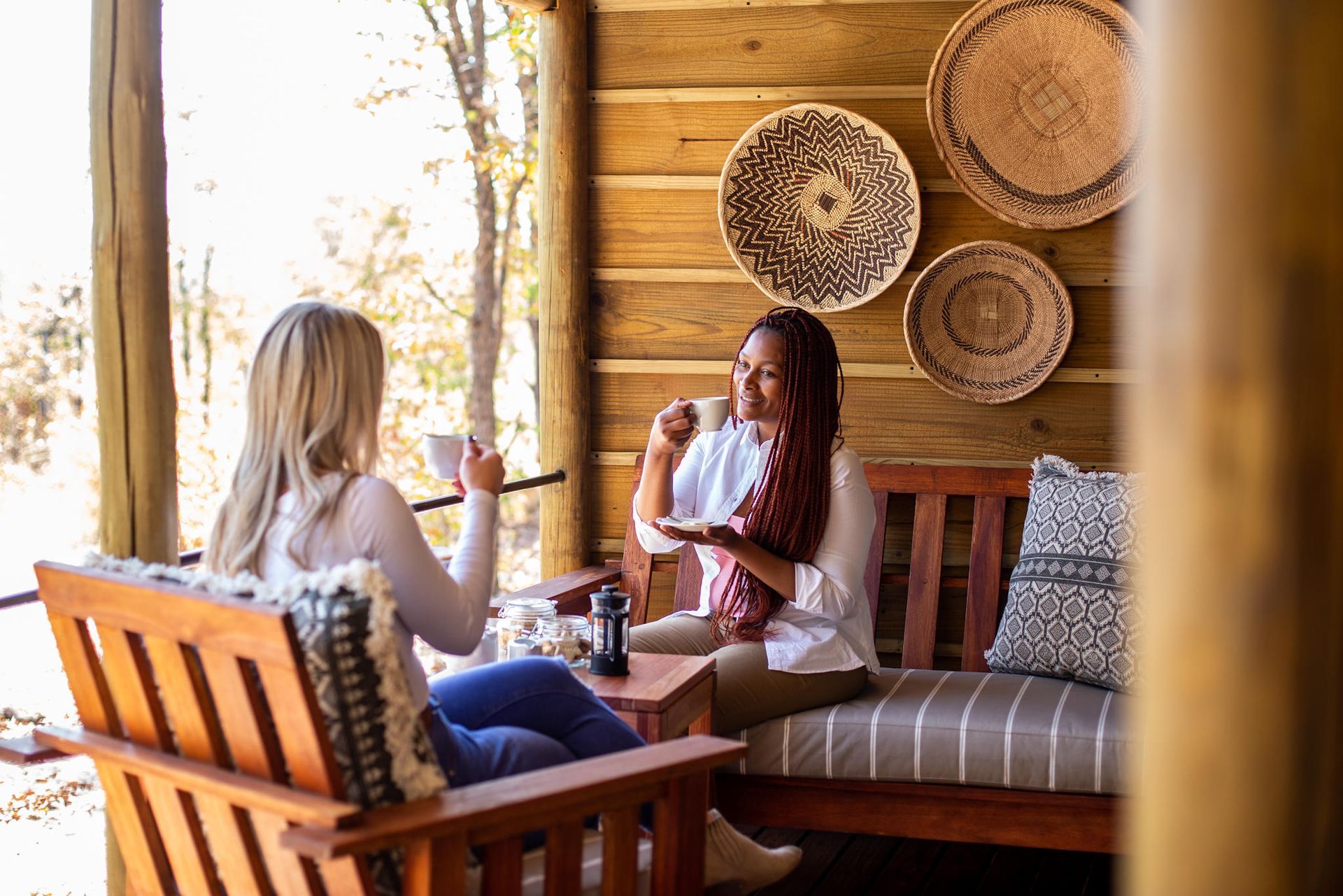 Two women drinking coffee on a wood porch, smiling. Baskets hang on the wall.