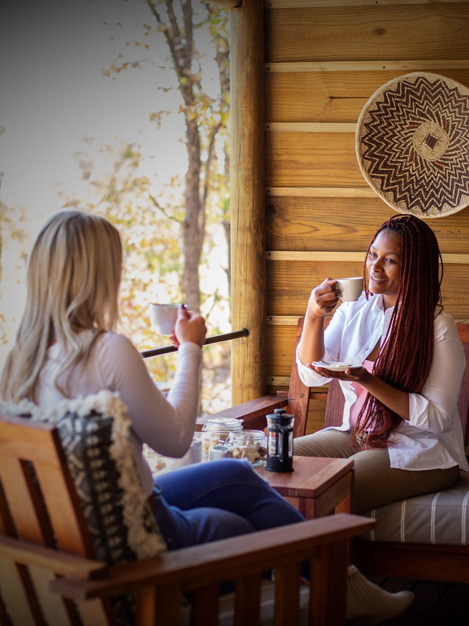 Two women drinking coffee on a wood porch, smiling. Baskets hang on the wall.