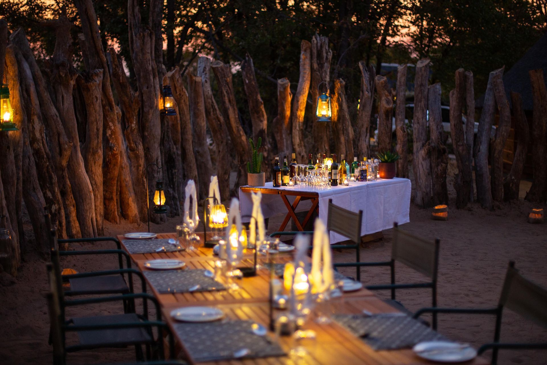 Outdoor dining table set with candles and lanterns, framed by a wooden fence, at dusk.