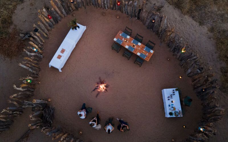 An outdoor dinner setting with tables, chairs, and a fire pit in a circular enclosure at dusk.