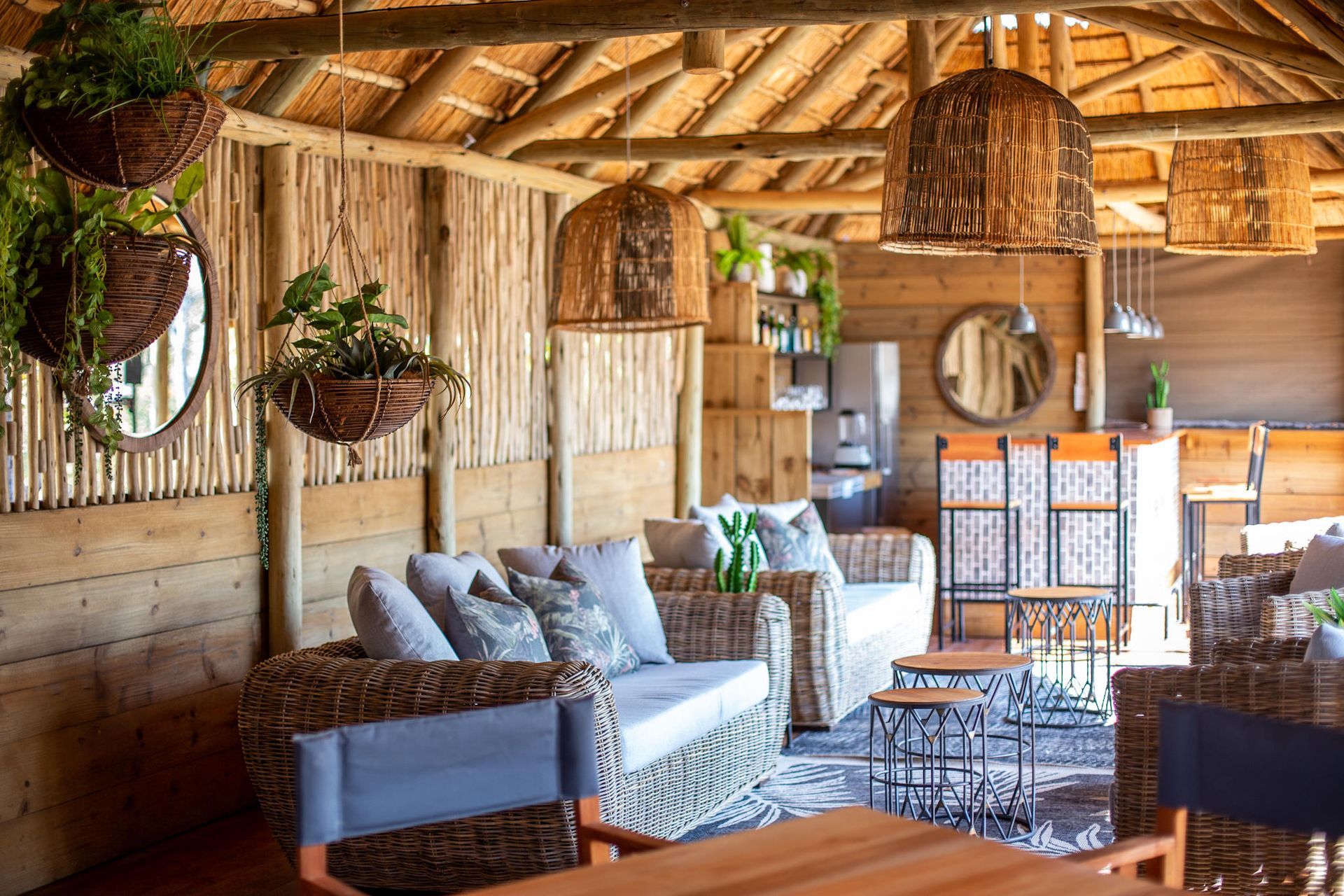 Cozy, rustic outdoor lounge with wicker furniture, hanging plants, and woven lamps under a thatched roof.