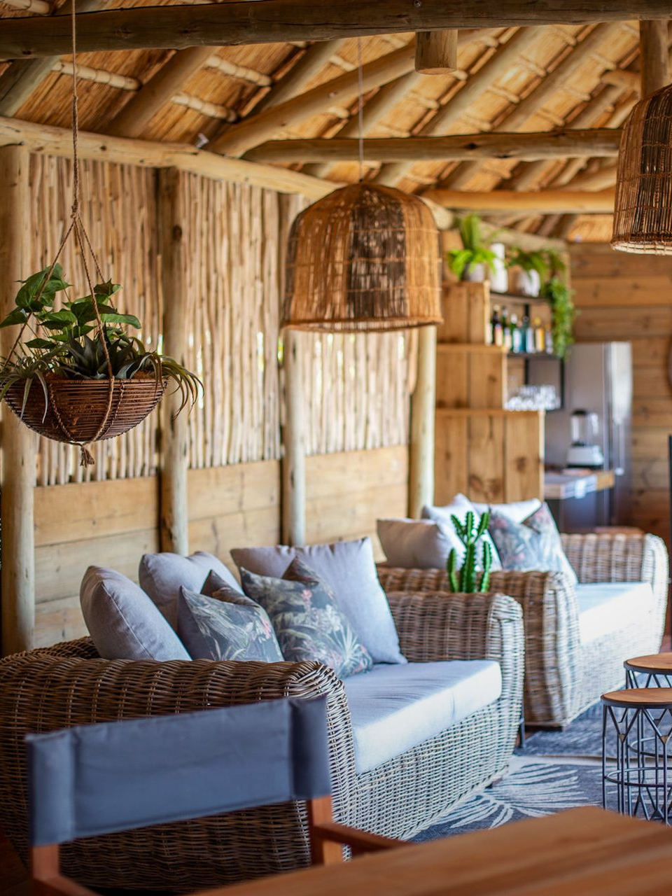 Cozy, rustic outdoor lounge with wicker furniture, hanging plants, and woven lamps under a thatched roof.