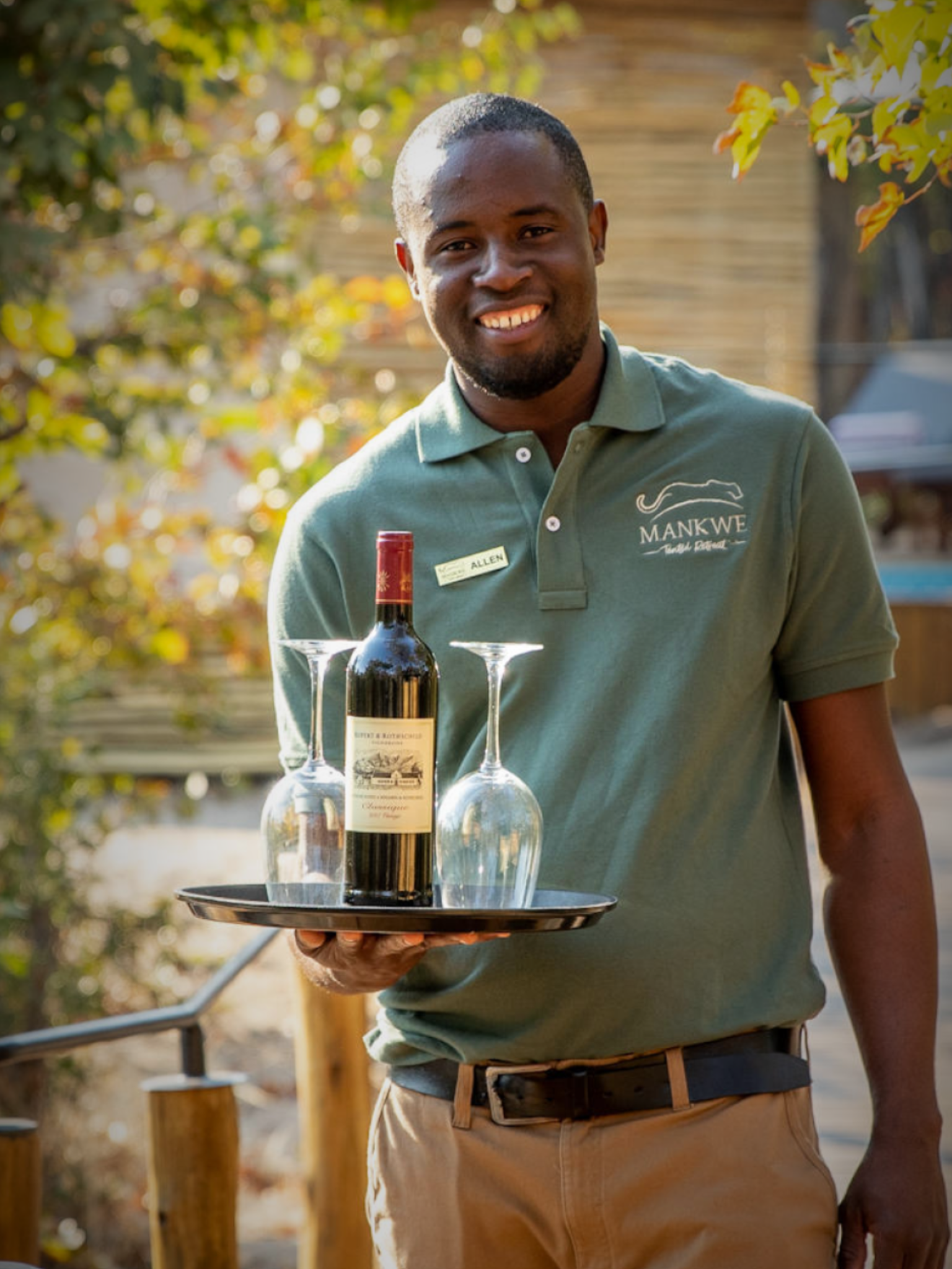 Waiter smiling, holding tray with wine bottle and glasses outdoors, wearing green shirt and khaki pants.