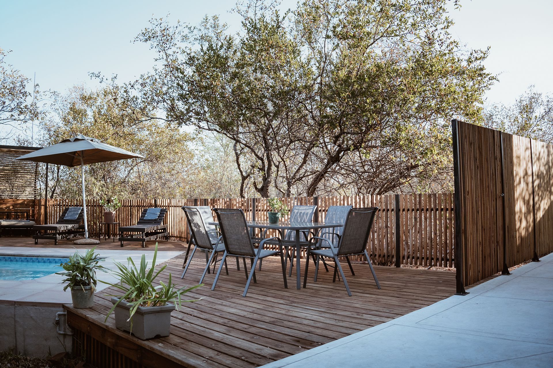 Wooden deck with outdoor dining table, chairs, pool, umbrella, and trees.