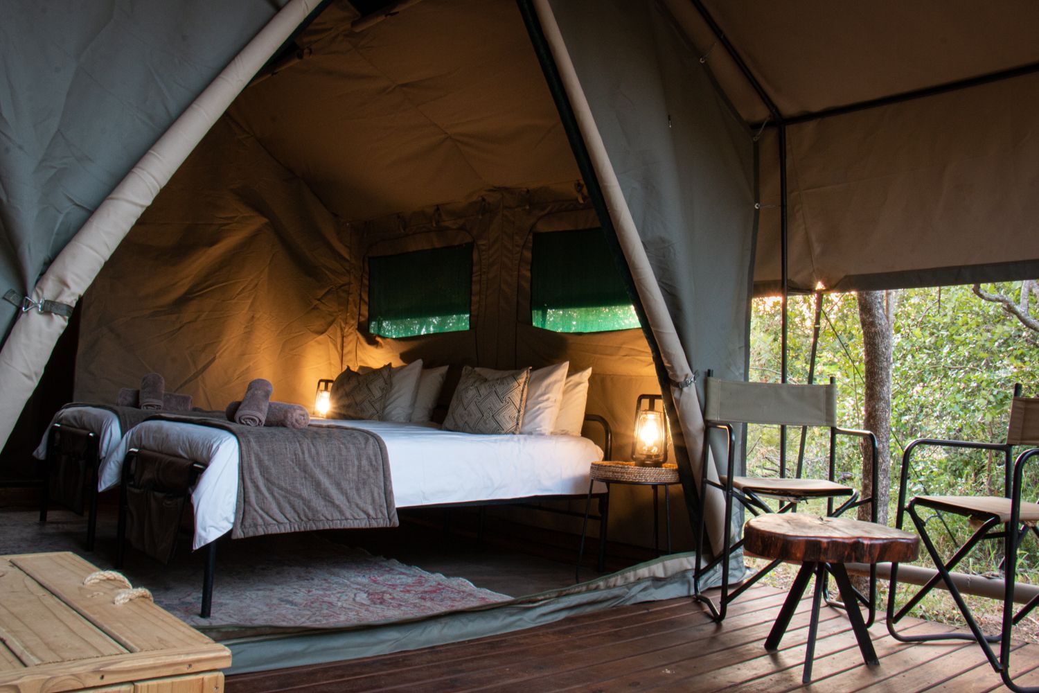 A luxury safari tent interior with a bed and seating on a wooden deck overlooking a forest.