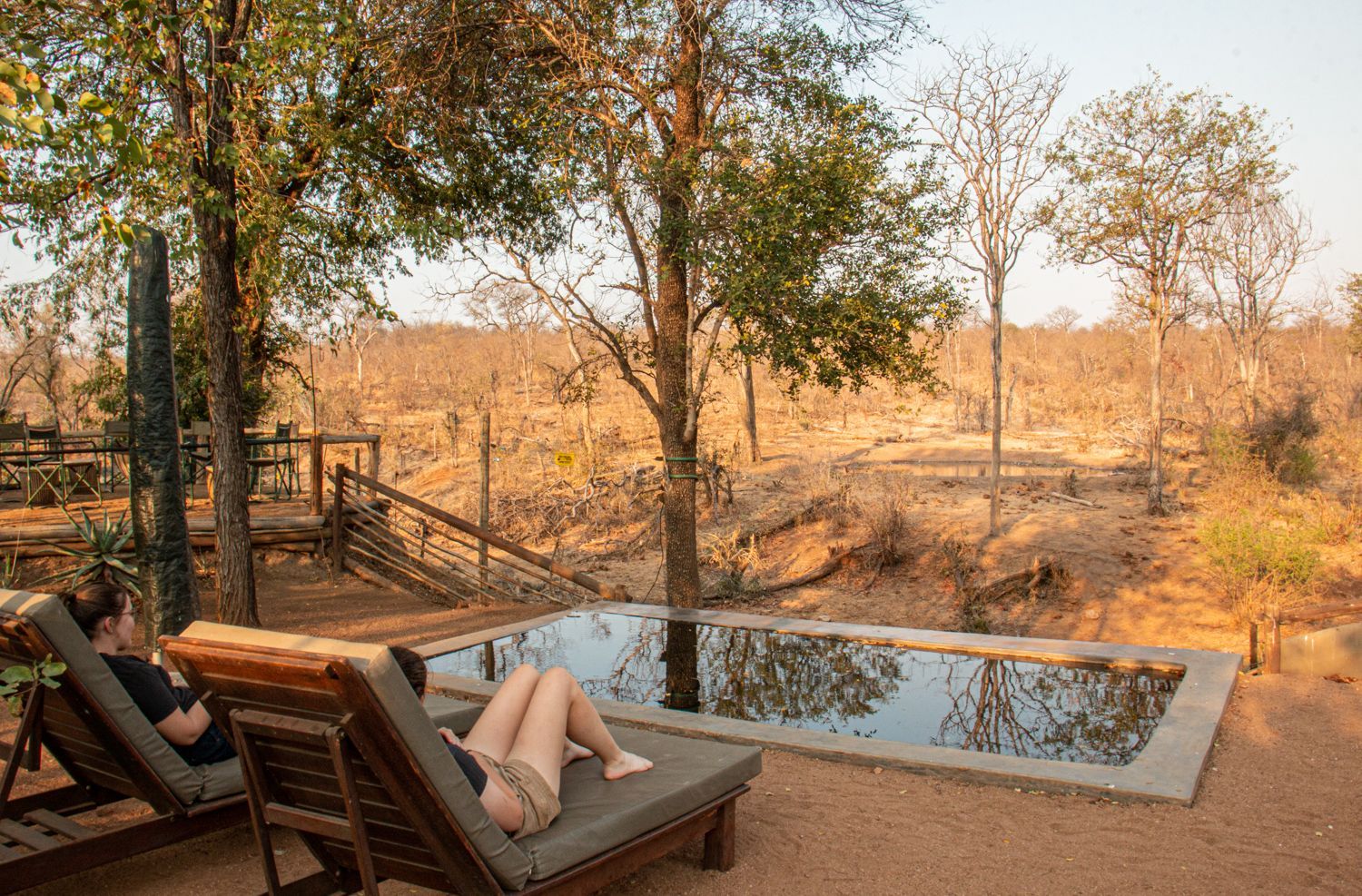 Two people lounging by a small infinity pool overlooking a dry, wooded landscape at sunset