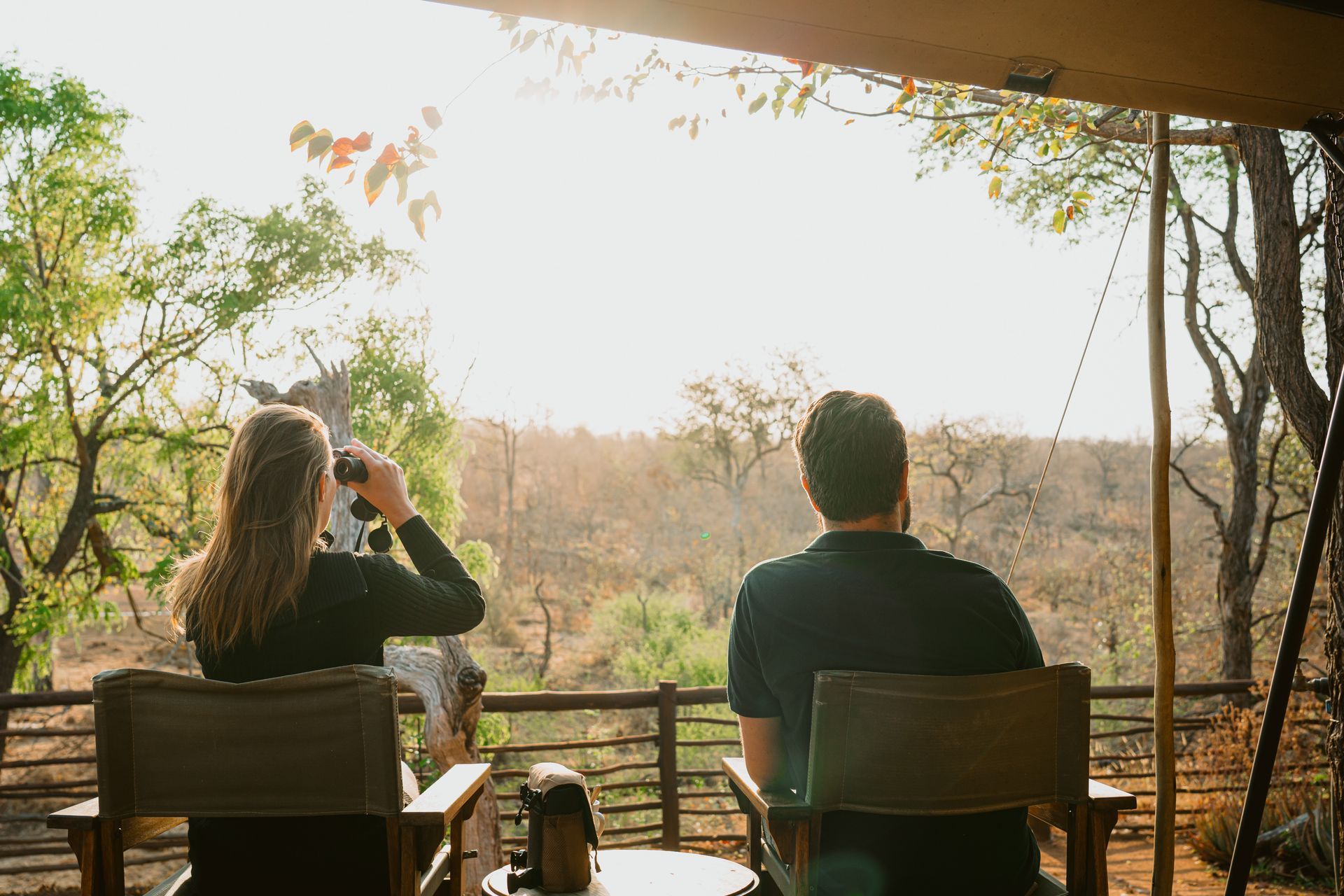Couple on a veranda overlooking a sunny landscape; woman using binoculars, man looking ahead.