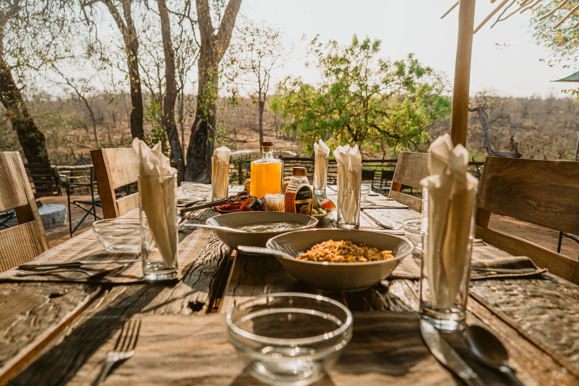 Outdoor dining table set with food and drinks; lush greenery visible in the background.