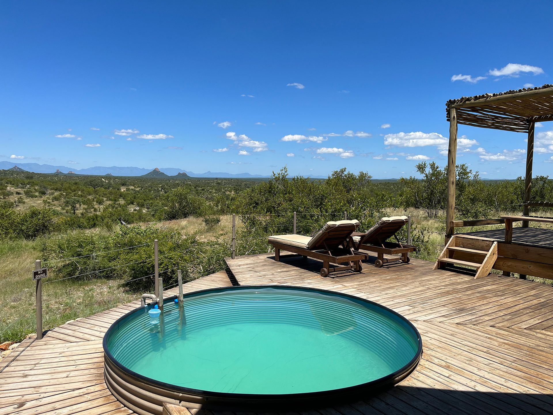 Round pool on a wooden deck with lounge chairs, overlooking a sunny landscape with blue sky.