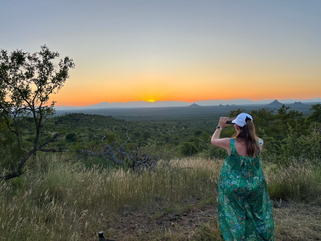 A person in a green dress and white cap stands on a grassy hill, using a phone to photograph a vibrant sunset over bush.