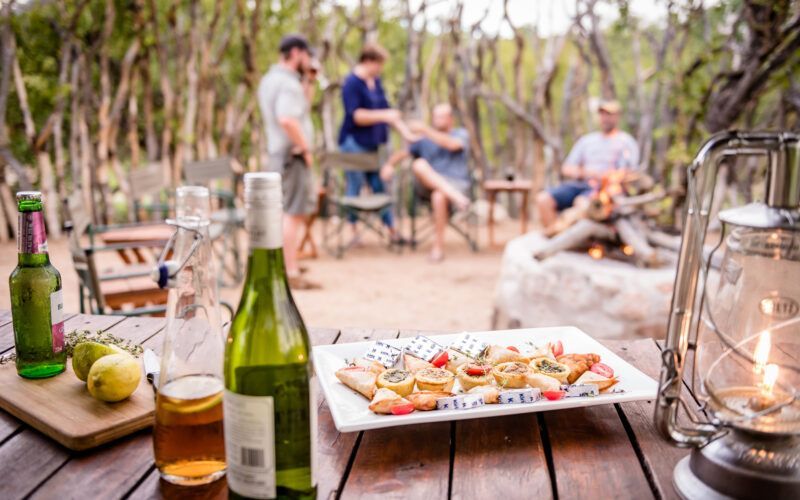 A picnic table with food and drinks in front of a campfire with people relaxing.