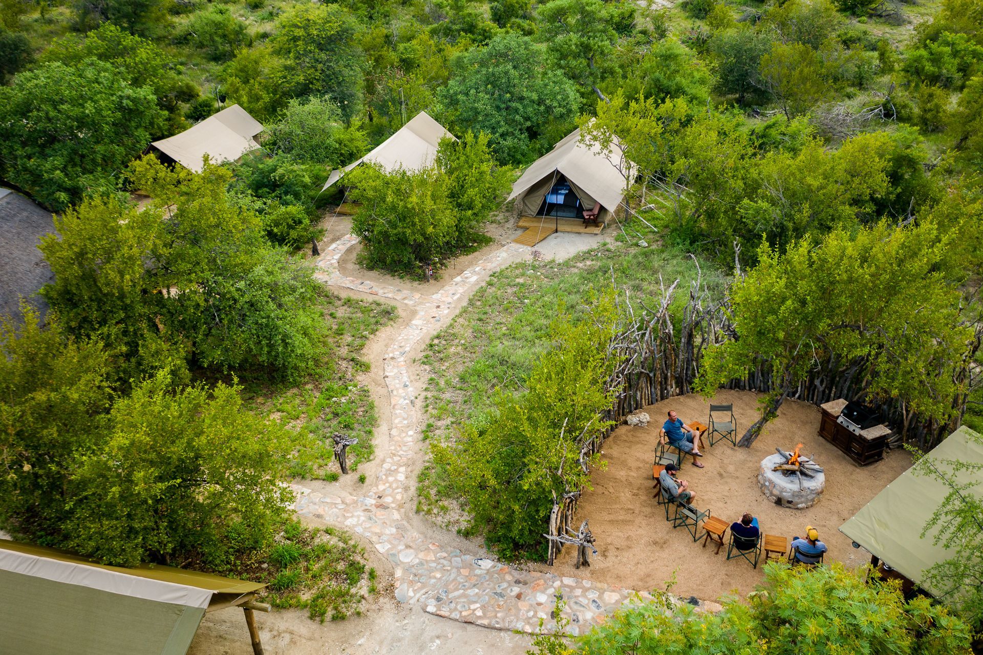 Safari camp with tents, surrounded by green foliage. Bundox guests sit around the fire pit