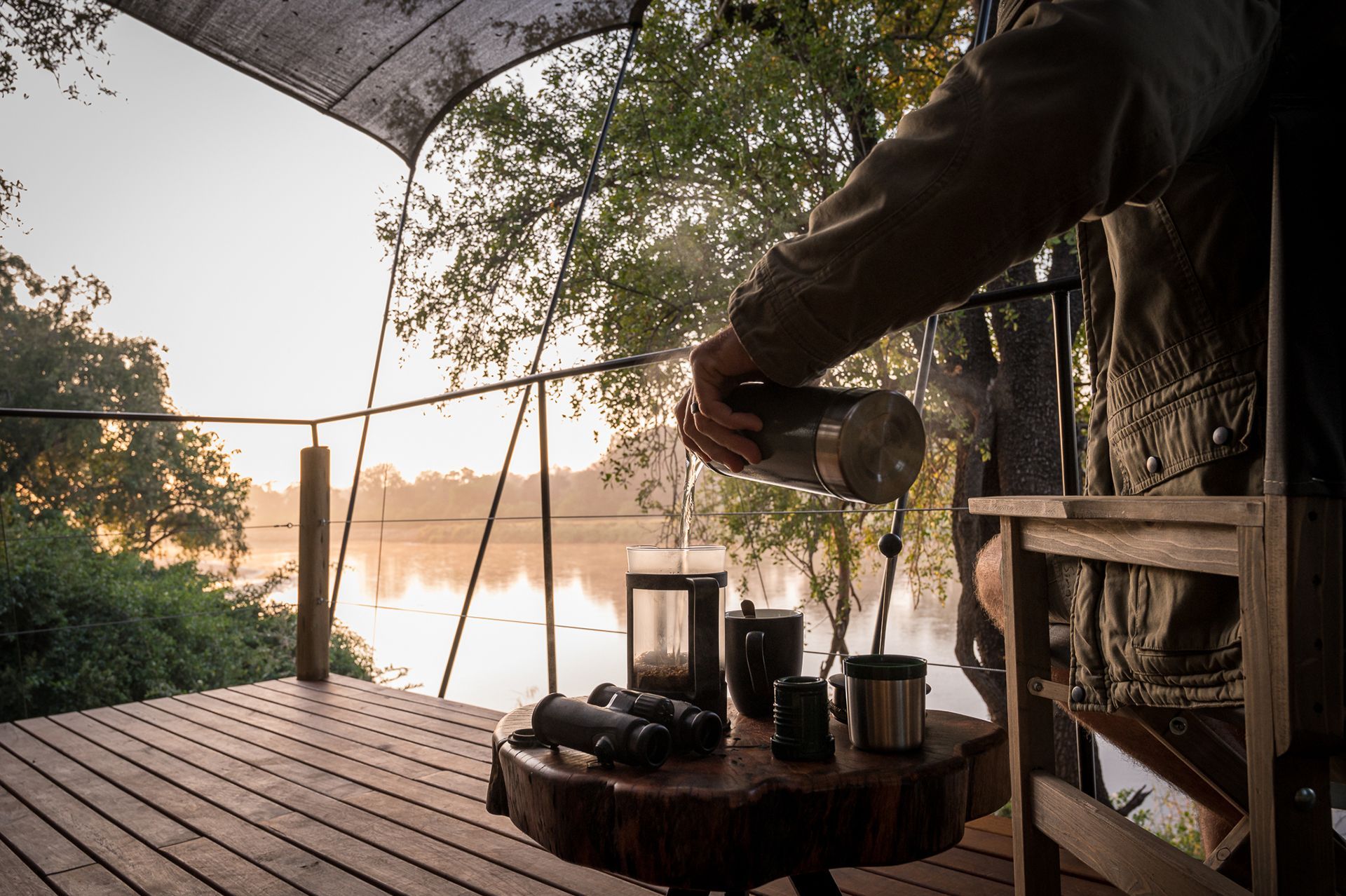 Bundox guests pouring liquid from a silver flask on a wooden deck overlooking a misty lake.