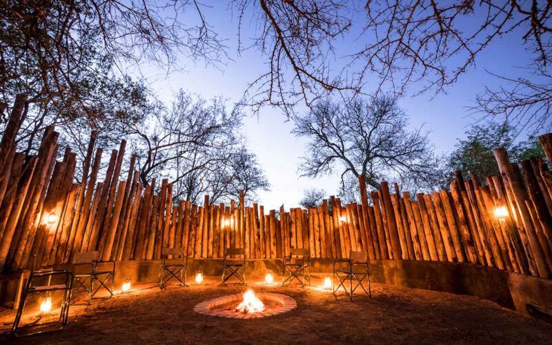 Campfire surrounded by a wooden fence with chairs and lanterns, trees in the background, dusk.