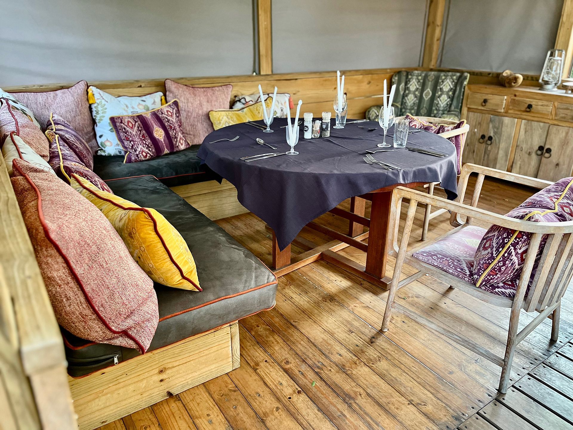 Cozy outdoor seating area with a round table set for a meal, surrounded by cushions and wooden furniture.