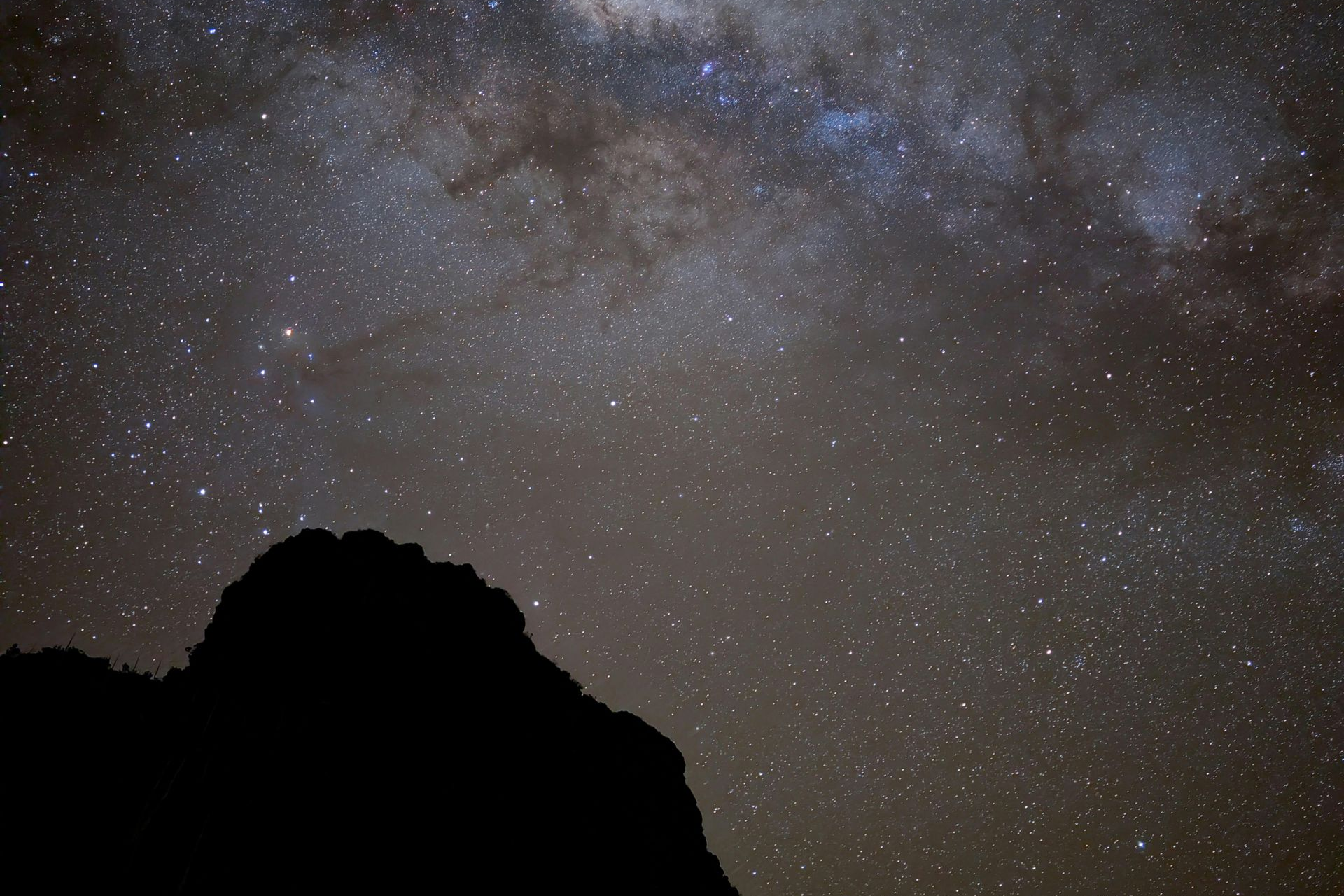 Starry night sky above a dark rocky silhouette, with the Milky Way visible overhead