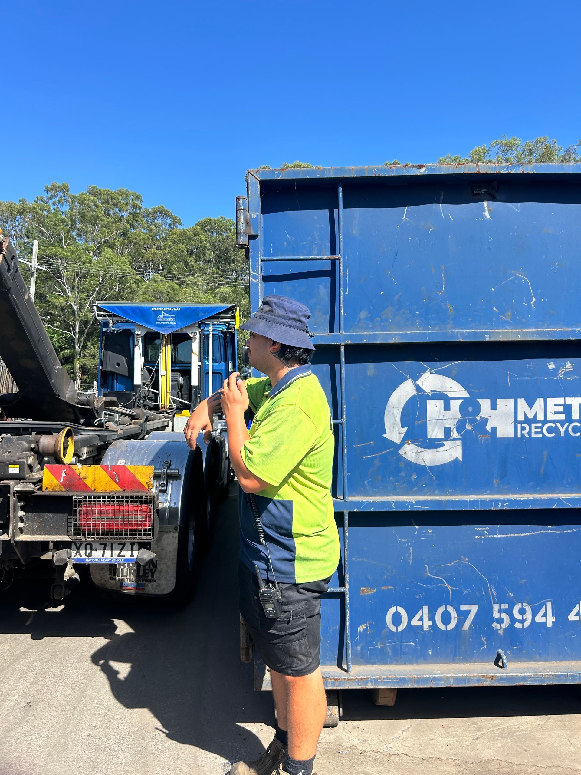 A Person in a High-visibility Lime Stands Beside a Blue Metal Recycling Skip — H&H Metal Recycling in Gympie, QLD