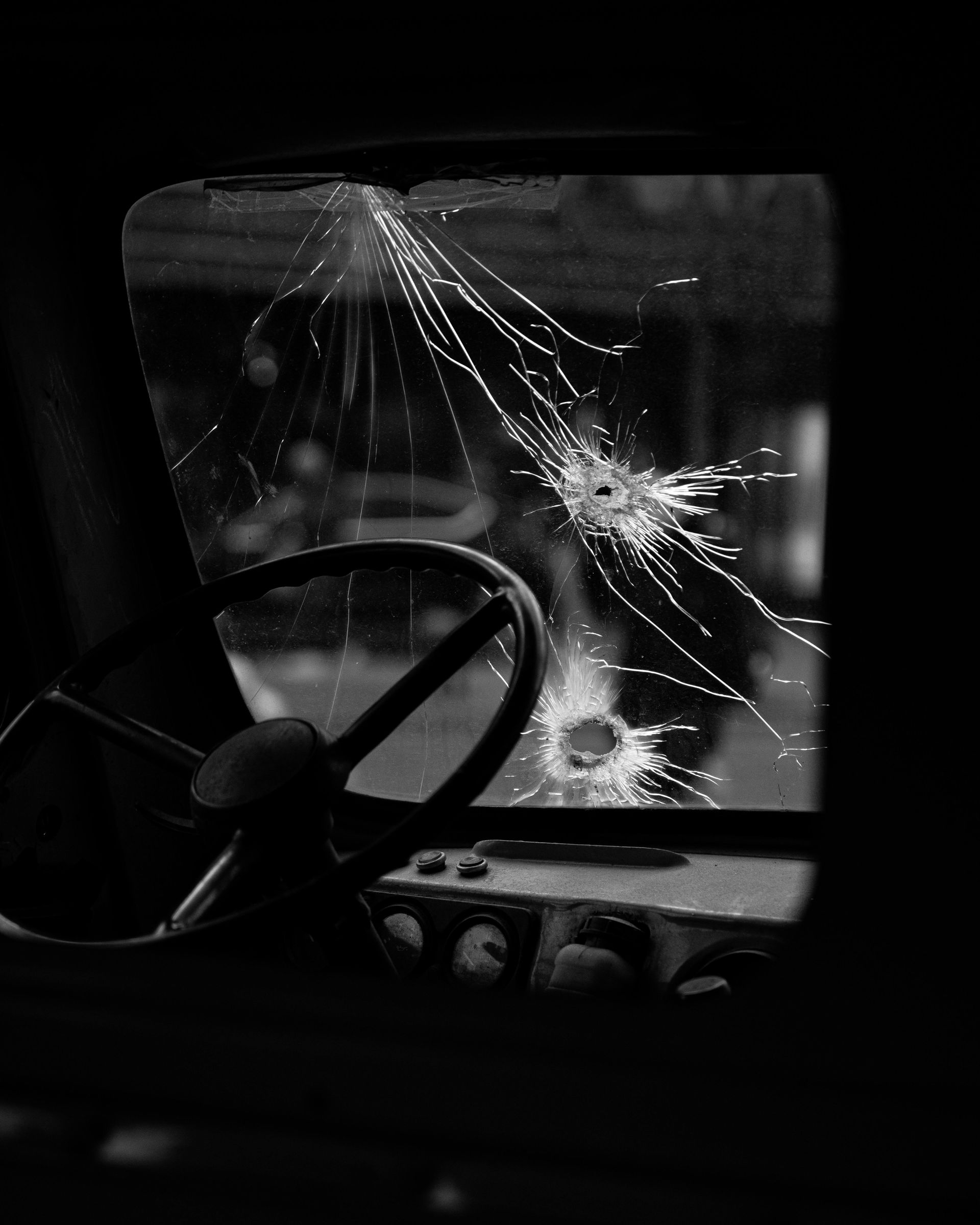 A black and white photo of a broken windshield with a steering wheel in the foreground.